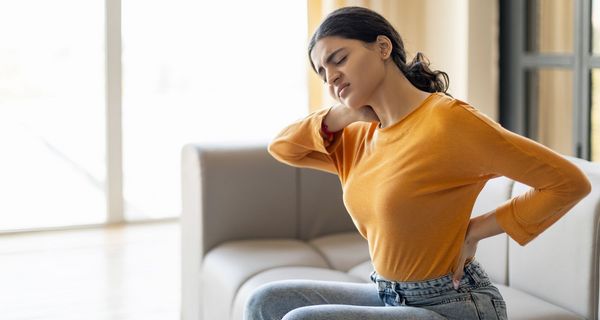 Junge Frau sitzt auf dem Sofa und hält sich den Rücken.