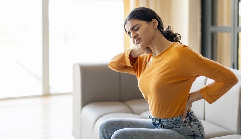 Junge Frau sitzt auf dem Sofa und hält sich den Rücken.