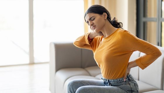 Junge Frau sitzt auf dem Sofa und hält sich den Rücken.