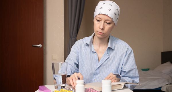 Eine Frau mit bedecktem Kopf sitzt in einem Krankenhauszimmer an einem Tisch. Vor ihr liegen zahlreiche Medikamentenblister und -dosen sowie ein aufgeschlagenes Buch.