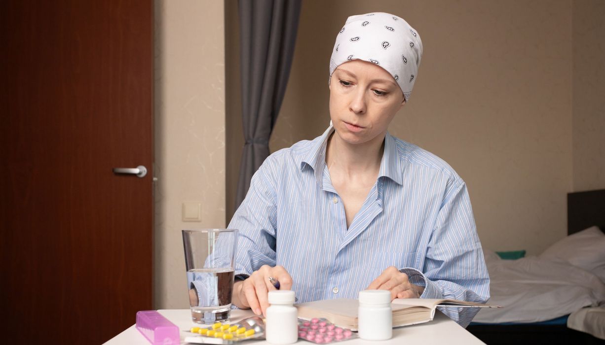 Eine Frau mit bedecktem Kopf sitzt in einem Krankenhauszimmer an einem Tisch. Vor ihr liegen zahlreiche Medikamentenblister und -dosen sowie ein aufgeschlagenes Buch.