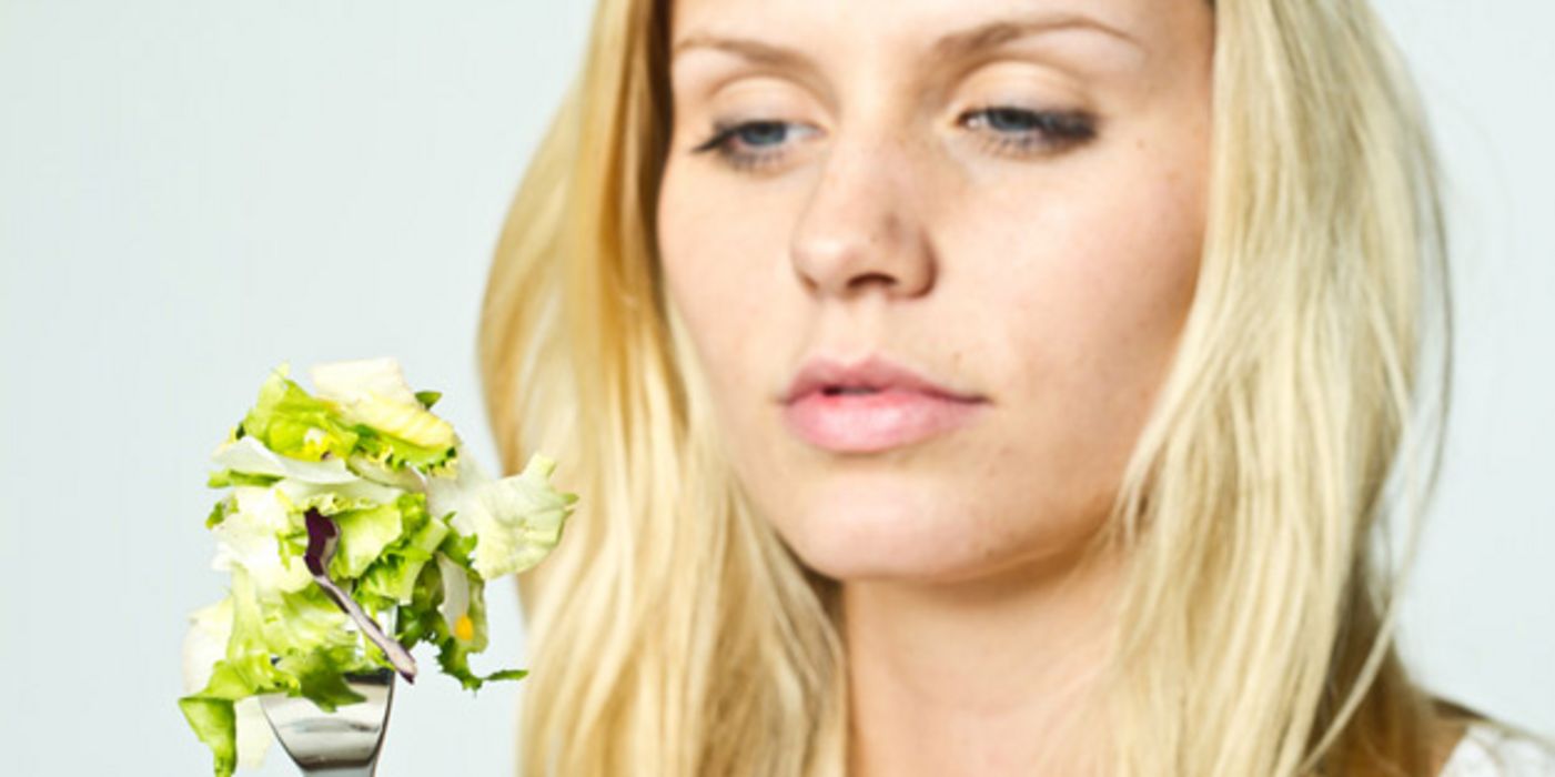 Nahaufnahme, blonde junge Frau, betrachtet mit verschlafenem Blick und angeödeter Miene ein Salatblatt, das sie auf eine Gabel gespießt hat