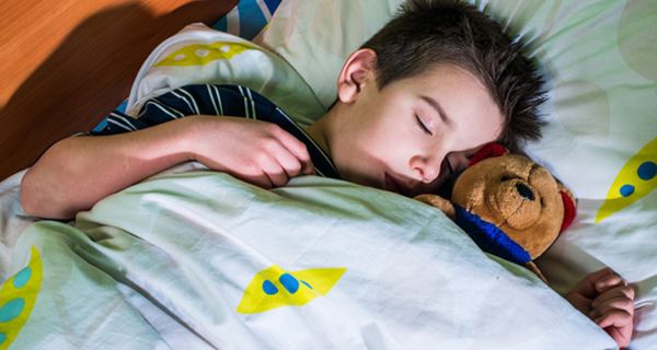 Schlafender Junge, ca. 8, mit Teddy im Arm, in buntem Bettzeug