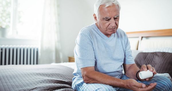 Mann, sitzt auf seinem Bett und schüttet eine Tablette aus einer Dose in seine Hand.