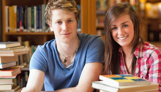 Student und Studentin in einer Bibliothek, umrundet von Bücherstapeln beim Lernen