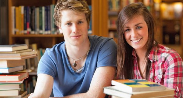 Student und Studentin in einer Bibliothek, umrundet von Bücherstapeln beim Lernen