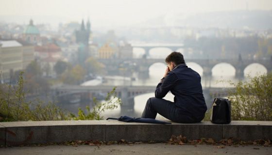 Rückansicht mit Halbprofil eines Mannes, auf einem Mäuerchen sitzend, das Gesicht in die Hände gestützt. Unter ihm die verhangene Stadtansicht von Prag und der Moldau