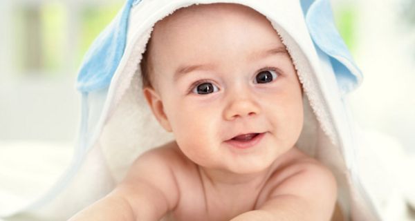 Porträt: Baby, auf dem Bauch liegend, weißhellblaues Handtuch auf dem Kopf, dunkle Augen, lächelnd, stützt sich auf Speckärmchen ab