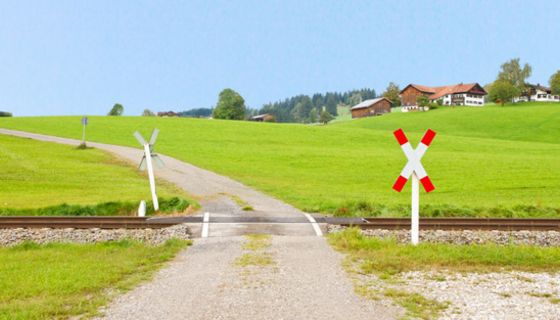 Bahnübergang auf dem Land, ein, zwei Häuser im Hintergrund, drumherum Wiesen