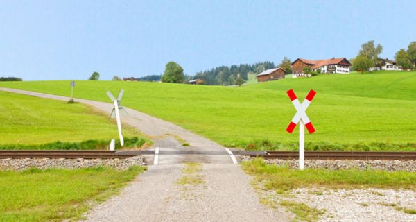 Bahnübergang auf dem Land, ein, zwei Häuser im Hintergrund, drumherum Wiesen