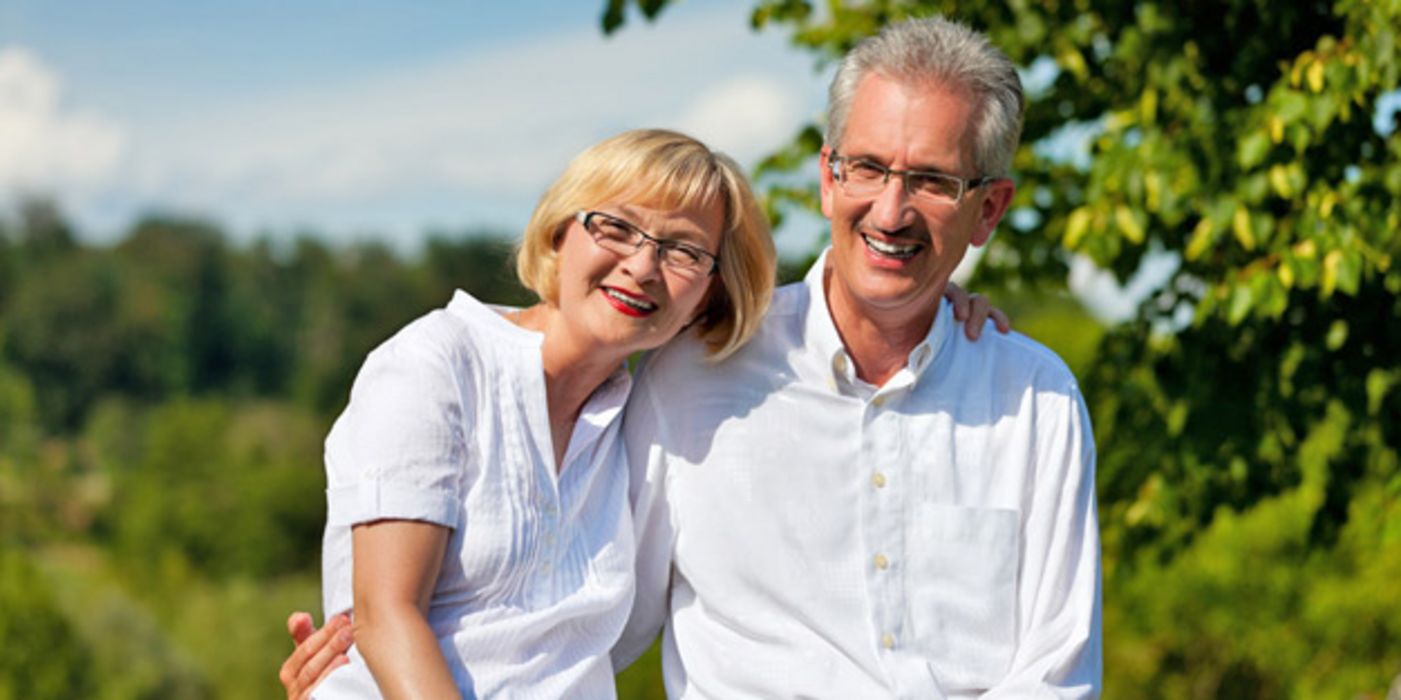 Mittelaltes Paar, weiße Bluse, weißes Hemd steht im sommergrünen Wald und lächelt in die Kamera