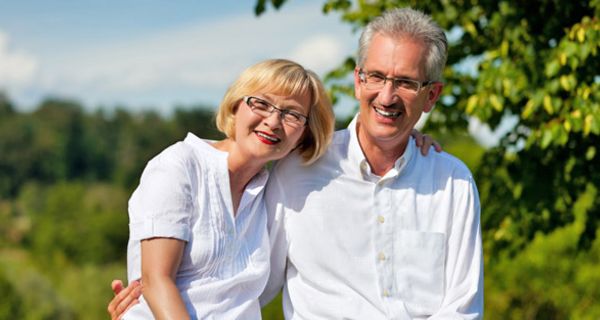 Mittelaltes Paar, weiße Bluse, weißes Hemd steht im sommergrünen Wald und lächelt in die Kamera