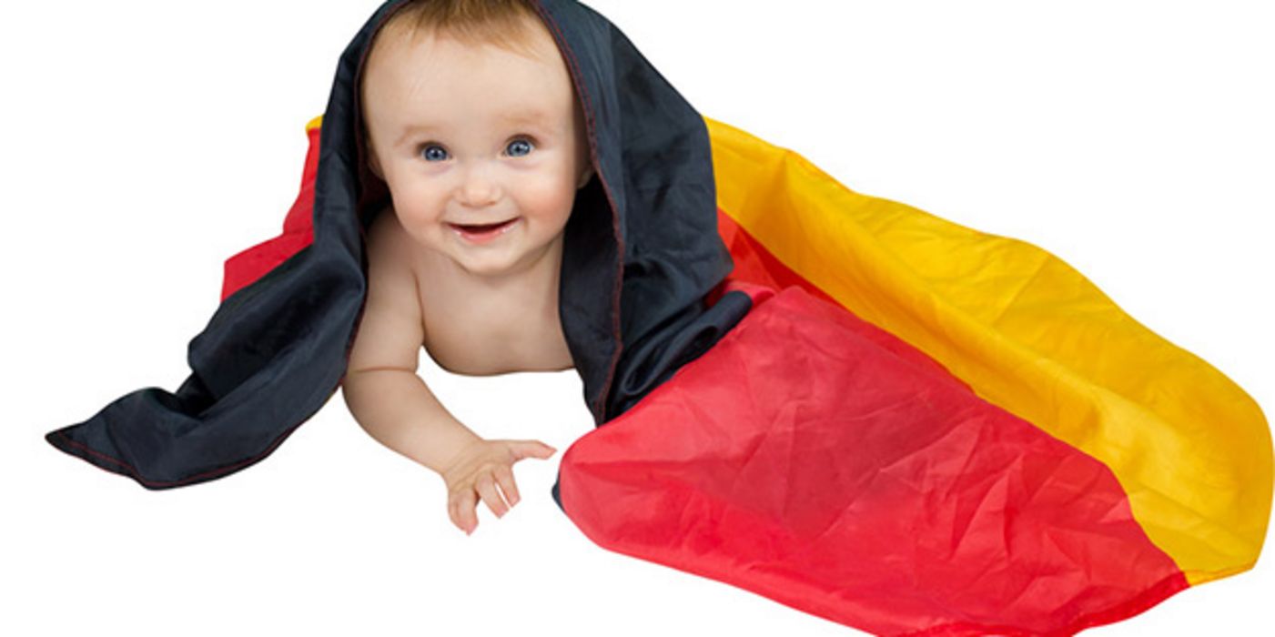 Freigestellt: Baby auf Bauch mit Deutschlandfahne über Kopf und auf Boden drapiert, lächelt in Kamera