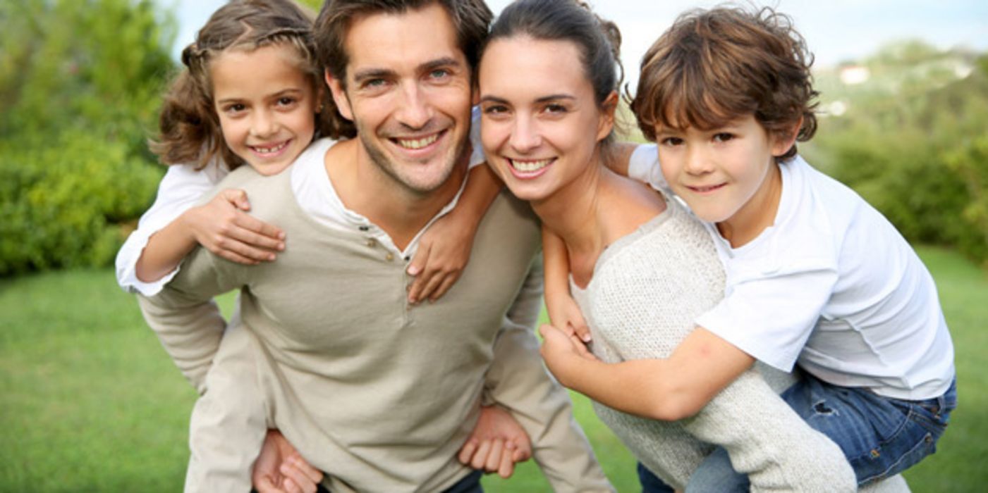 Attraktive Familie mit 2 Kindern auf Rasen huckepack (Junge bei Mutter, Mädchen bei Vater) lachen in die Kamera