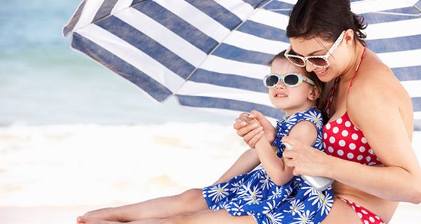 Mutter mit Kind in bunter Badekleidung (blau, weiß, rot) unter blau-weißem Sonnenschirm. Mädchen sitzt auf Mutters Beinen, wird eingecremt