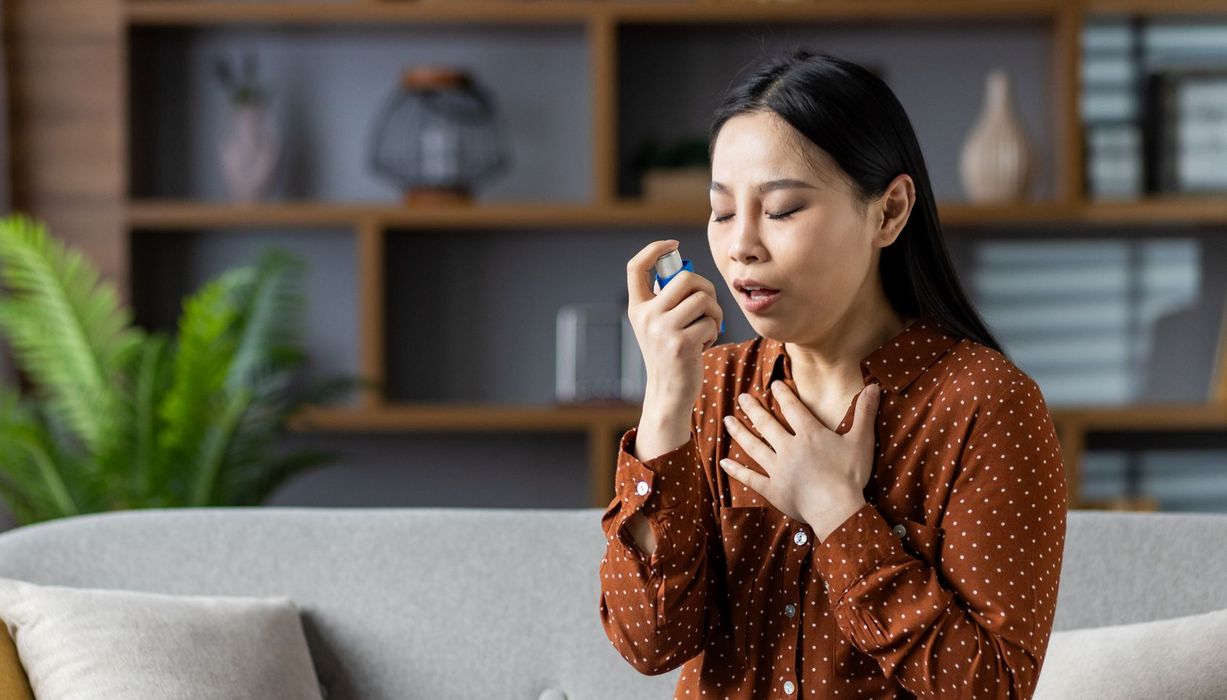 Frau sitzt auf dem Sofa und verwendet einen Inhalator. Eine Hand liegt an ihrem Schlüsselbein.