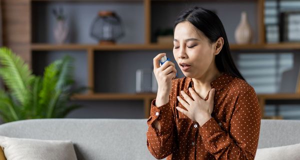 Frau sitzt auf dem Sofa und verwendet einen Inhalator. Eine Hand liegt an ihrem Schlüsselbein.