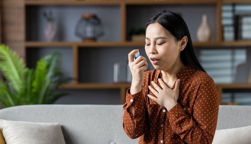 Frau sitzt auf dem Sofa und verwendet einen Inhalator. Eine Hand liegt an ihrem Schlüsselbein.