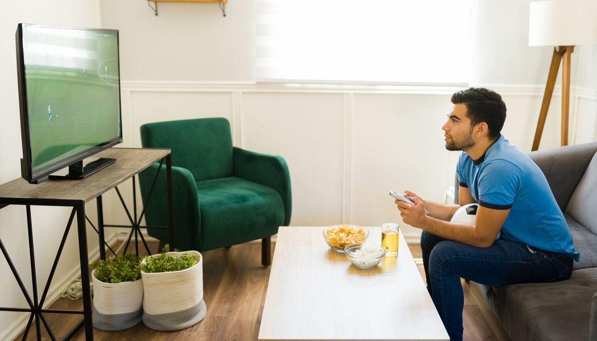 Junger Mann schaut ein Fußballspiel auf dem Fernseher. Auf dem Schoß hat er einen Fußball. Tageslicht fällt durch ein Fenster.