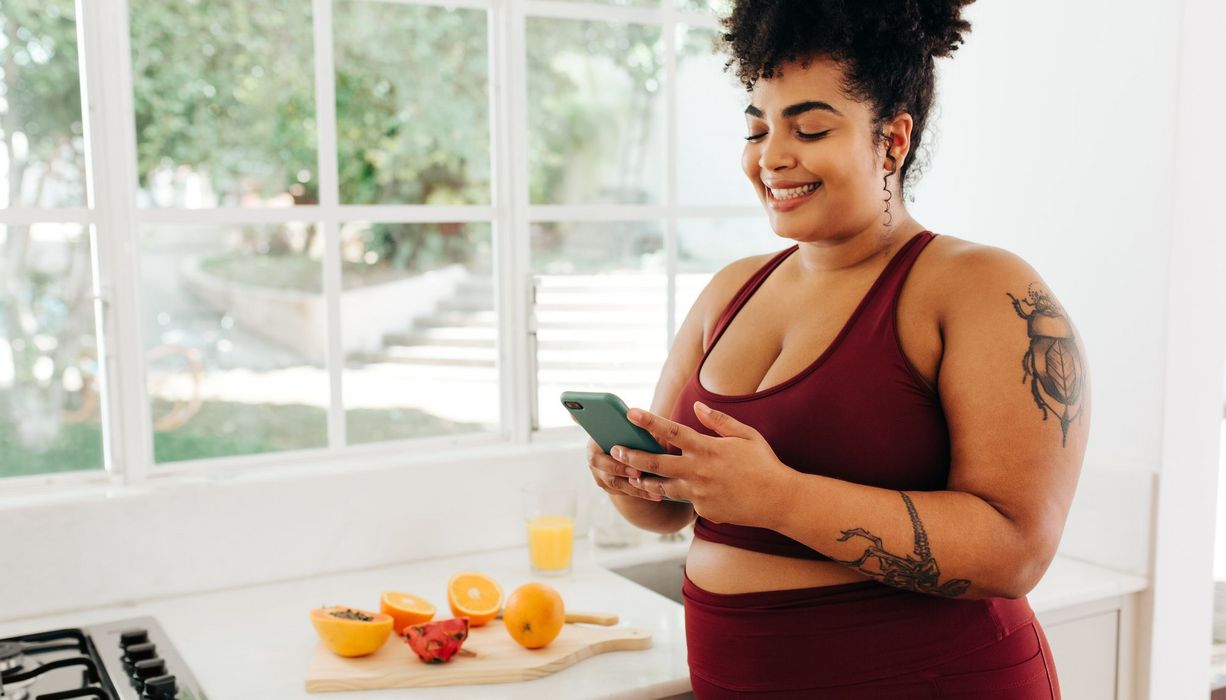 Junge Frau in Sportkleidung mit Übergewicht schaut lächelnd auf ihr Smartphone. AUf der Küchenanrichte liegen aufgeschnittene Früchte.