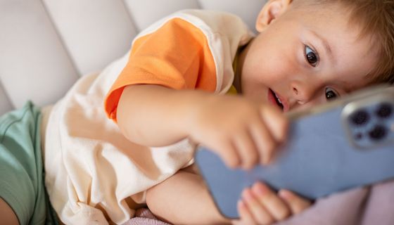 Kleinkind liegt mit einem Smartphone in den Händen auf dem Sofa und blickt darauf.