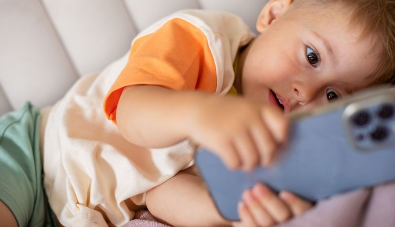 Kleinkind liegt mit einem Smartphone in den Händen auf dem Sofa und blickt darauf.