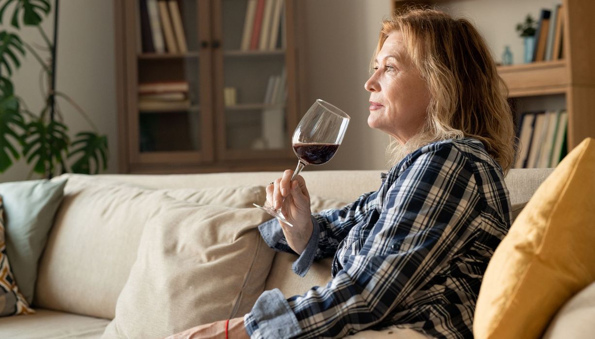 Eine mittelalte Frau sitzt auf dem Sofa und trinkt allein ein Glas Wein.