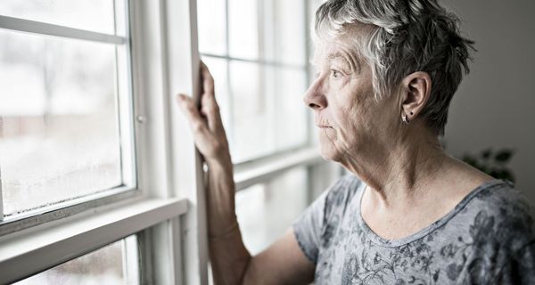 Ältere Dame, steht am Fenster und schaut traurig nach draußen.