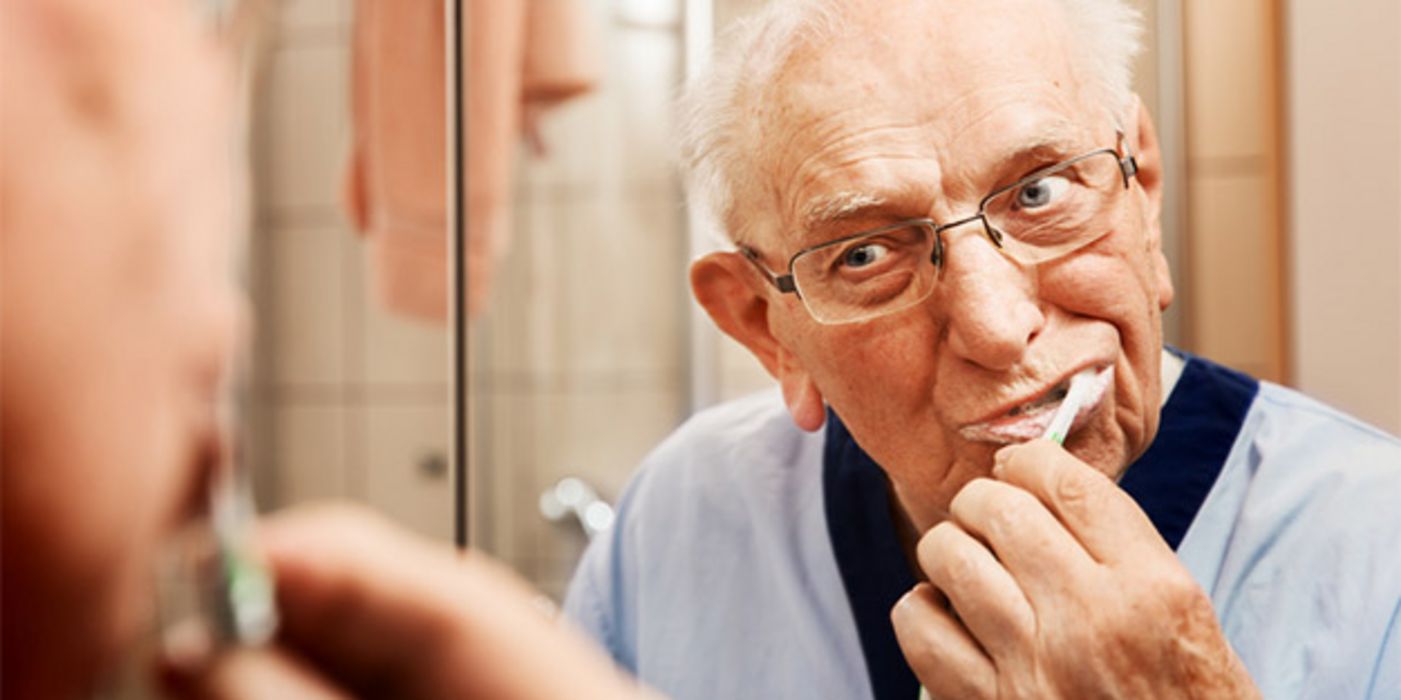 Alter Mann mit Brille putzt sich die Zähne; im Spiegel zu sehen