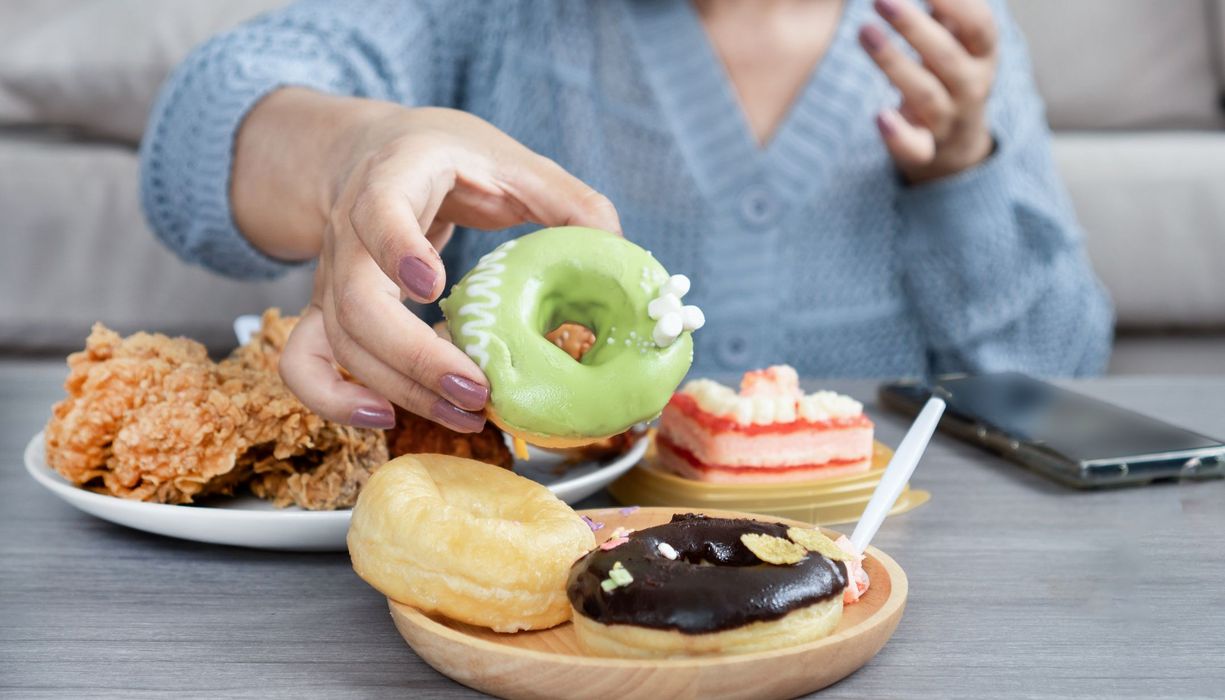 Frau vor mehreren Tellern mit Donuts, Fast Food und Kuchen.