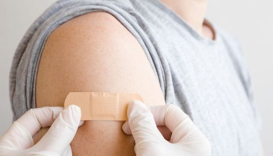 Zwei Hände in Handschuhen kleben ein Pflaster auf den Oberarm eines Geimpften.