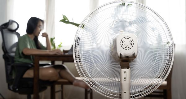 Vordergrund: Ein eingeschalteter Ventilator von hinten, im Hintergrund unscharf: Eine junge Frau arbeitet an einem Laptop.
