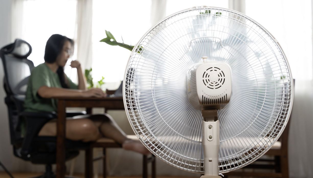 Vordergrund: Ein eingeschalteter Ventilator von hinten, im Hintergrund unscharf: Eine junge Frau arbeitet an einem Laptop.