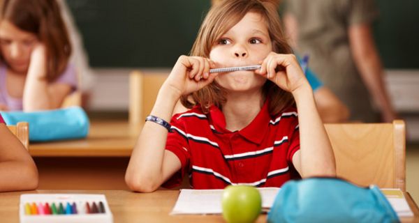 Grundschüler sitzt am Pult, hat einen Bleistift zwischen den Lippen und denkt nach