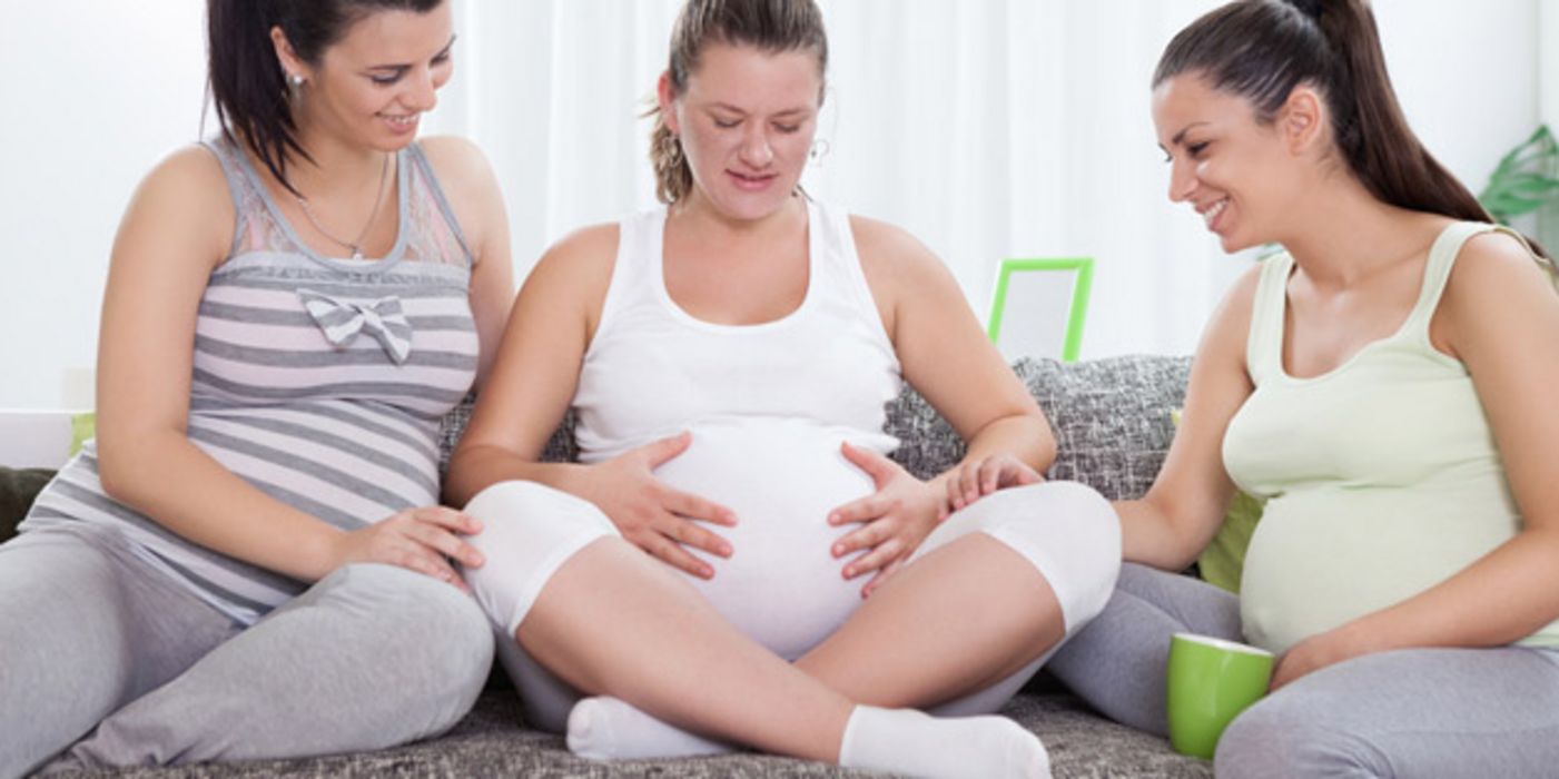 Drei hoch schwangere Frauen sitzen nebeneinander auf einer Couch und begutachten den Bauch der in der Mitte Sitzenden