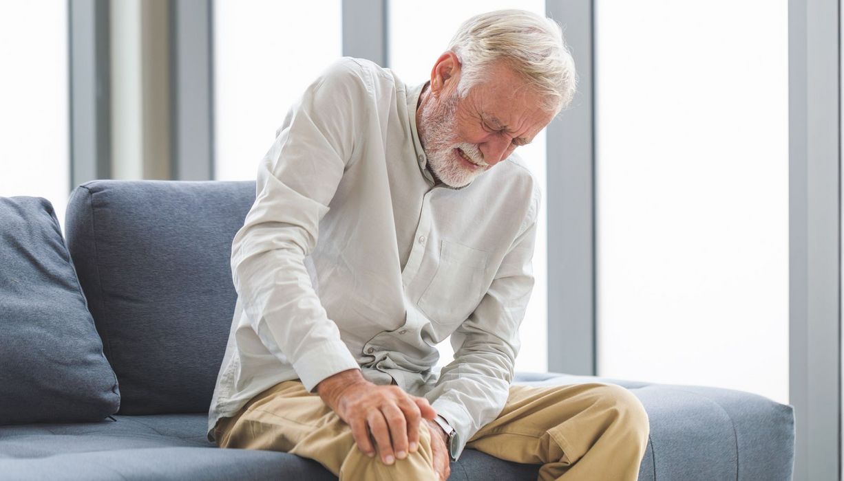Ein älterer Mann sitzt mit schmerzverzerrtem Gesicht auf dem Sofa und hält sich sein Knie.