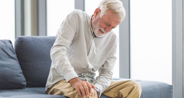 Ein älterer Mann sitzt mit schmerzverzerrtem Gesicht auf dem Sofa und hält sich sein Knie.