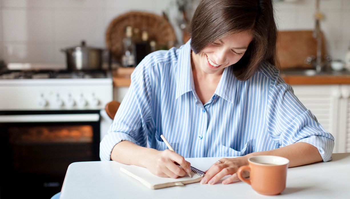 Junge Frau mit brünettem Haar sitzt am Küchentisch und schreibt in ein Journal, lächelnd.