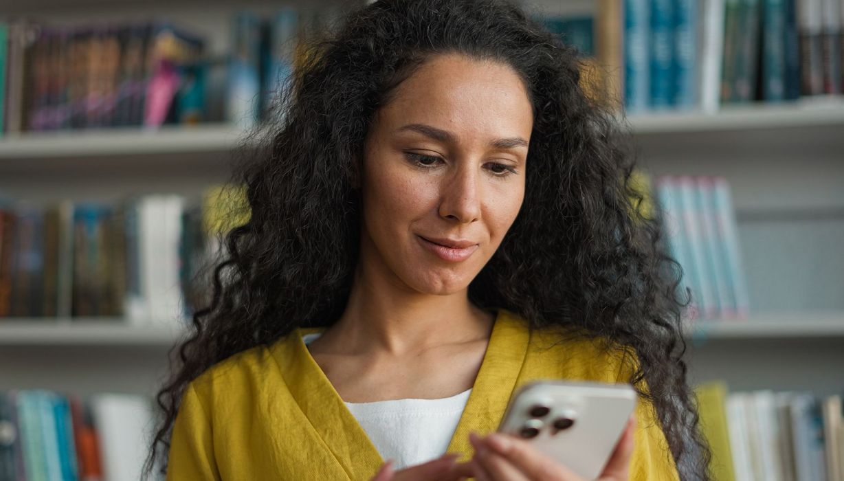 Junge Frau schaut auf ihr Smartphone, unscharf im Hintergrund ein Bücherregal.