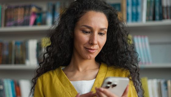Junge Frau schaut auf ihr Smartphone, unscharf im Hintergrund ein Bücherregal.