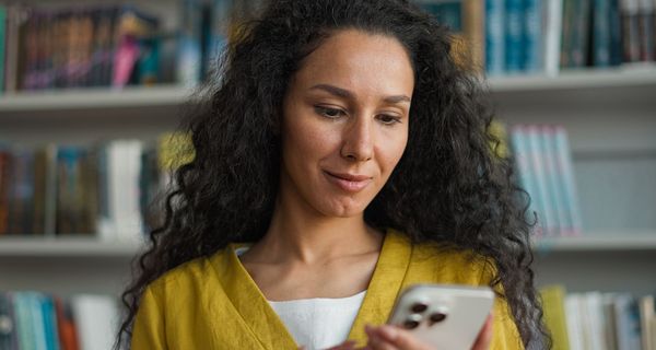 Junge Frau schaut auf ihr Smartphone, unscharf im Hintergrund ein Bücherregal.