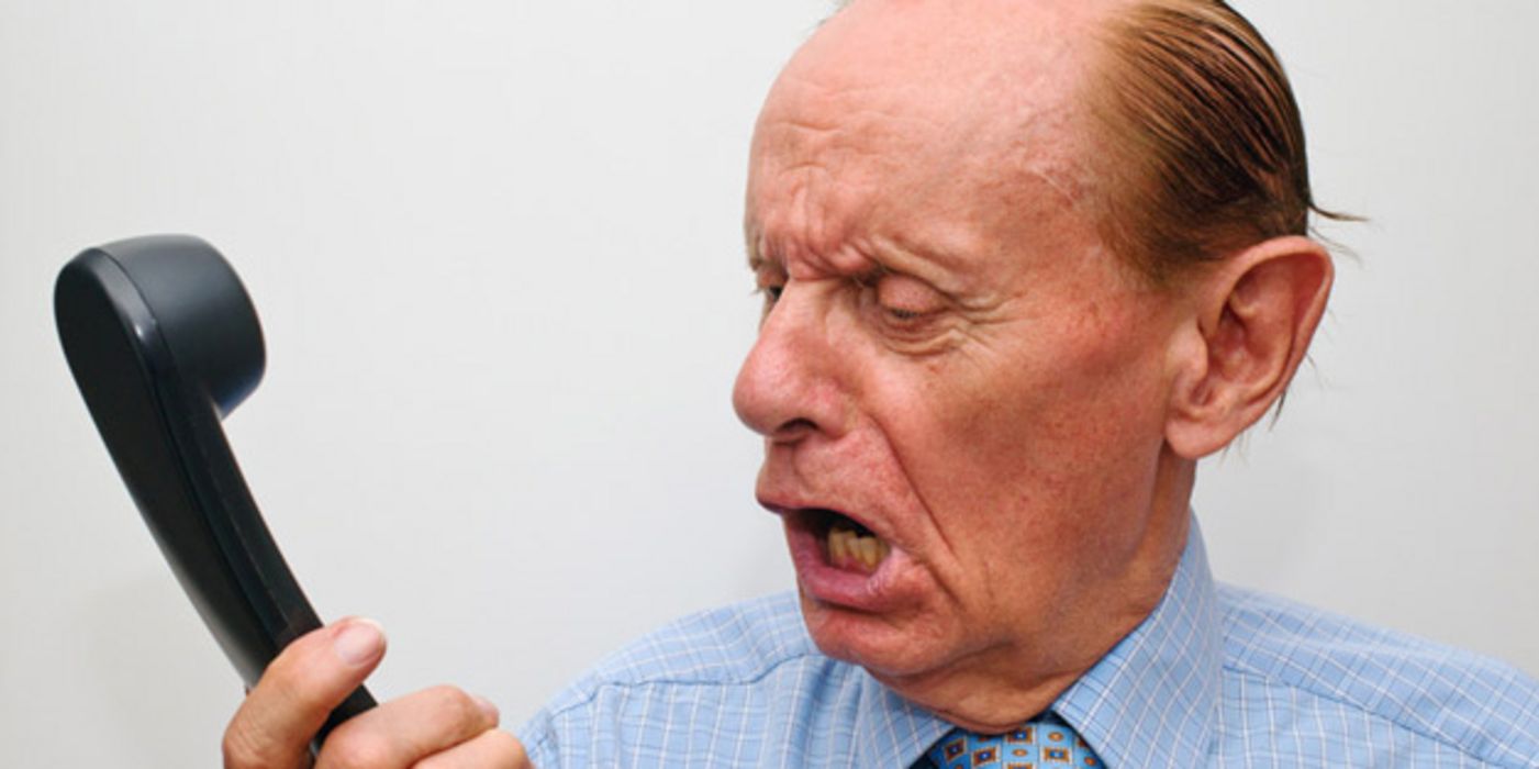 Älterer Mann in blauem Hemd und Schlips schaut wütend auf einen Telefonhörer in seiner Hand und brüllt hinein