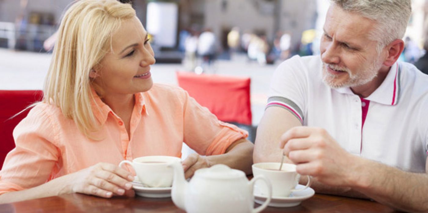 Älteres Paar, einander zugewandt, lächelnd (er ca. 60+, sie etwas jünger), sitzen im Straßencafé und unterhalten sich