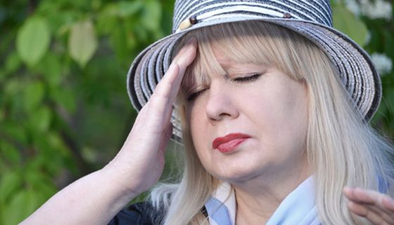 Im Freien (Sträucher oder Bäume im Hintergrund): Frau ca. Mitte 50 mit Sommerstrohhut (blau-weiß), geschlossene Augen, greift sich mit der rechten Hand an die schmerzende Stirn.