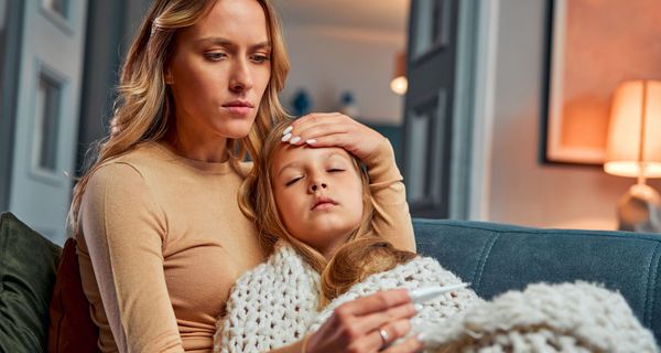Krankes Kind liegt im Arm einer Frau, eingehüllt in eine Decke. Die Frau hat eine Hand an die Stirn des Kindes gelegt und liest ein Fieberthermometer ab.