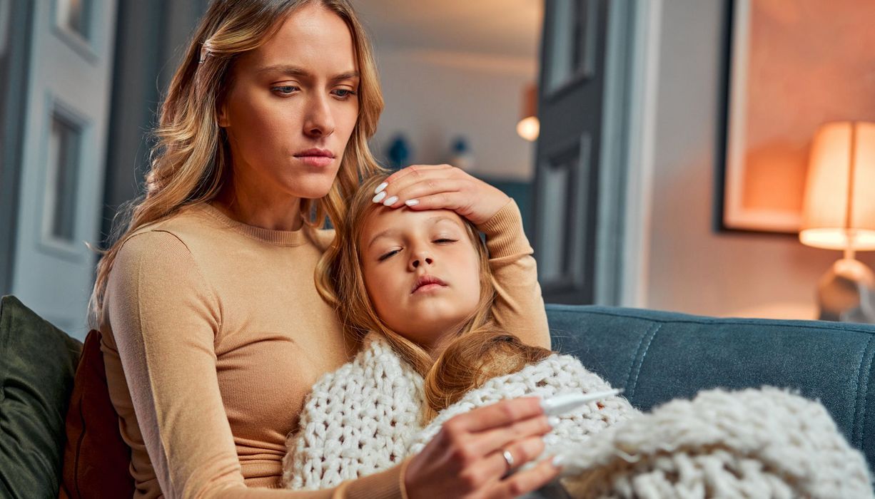 Krankes Kind liegt im Arm einer Frau, eingehüllt in eine Decke. Die Frau hat eine Hand an die Stirn des Kindes gelegt und liest ein Fieberthermometer ab.