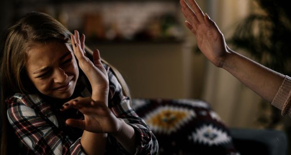 Mädchen duckt sich und hebt abwehrend die Hände – am rechten Bildrand ist eine drohend erhobene Hand zu sehen.