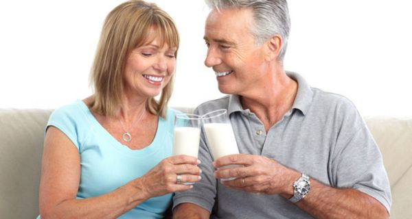 Attraktives Paar Ende 50, lachend, auf heller Couch sitzend, prostet sich mit Milch zu