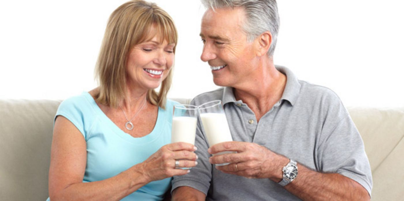 Attraktives Paar Ende 50, lachend, auf heller Couch sitzend, prostet sich mit Milch zu