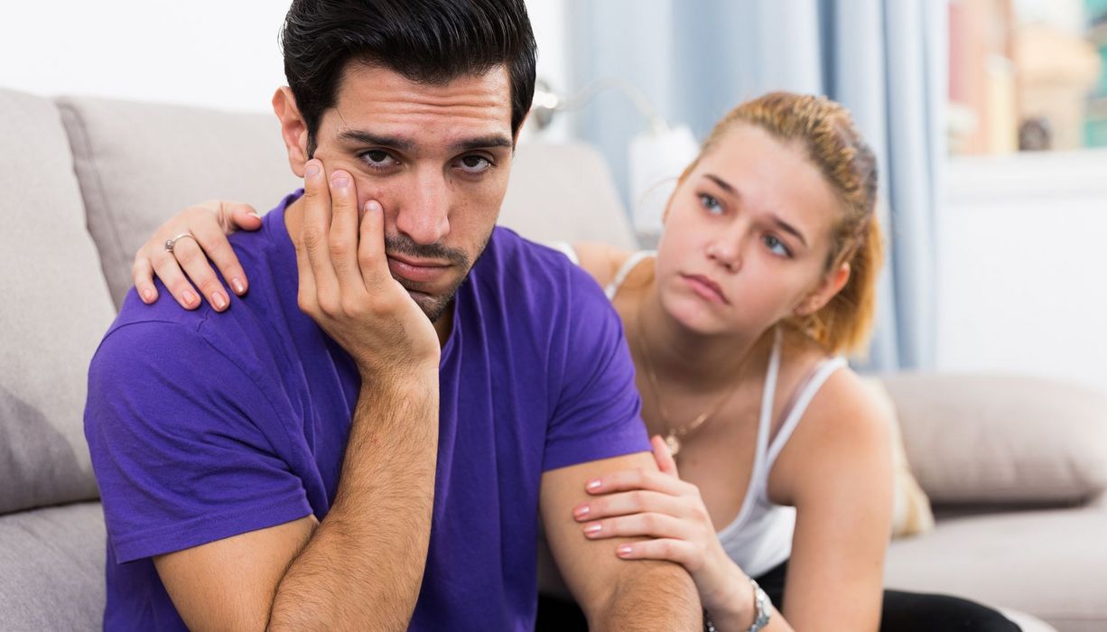 Mann sitzt auf einem Sofa und blickt unglücklich in die Kamera. Eine Frau hat ihm eine Hand auf die Schulter gelegt und versucht, ihm zu helfen.